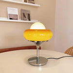 Load image into Gallery viewer, Crostata Table Lamp with a polished chrome base and vibrant orange glass bowl, placed on a white tabletop in a minimalist room, accented by bookshelves and neutral decor for a modern and cozy ambiance.
