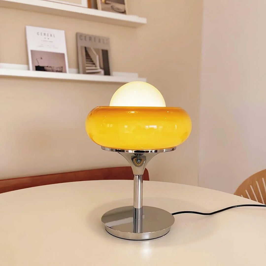 Crostata Table Lamp with a polished chrome base and vibrant orange glass bowl, placed on a white tabletop in a minimalist room, accented by bookshelves and neutral decor for a modern and cozy ambiance.