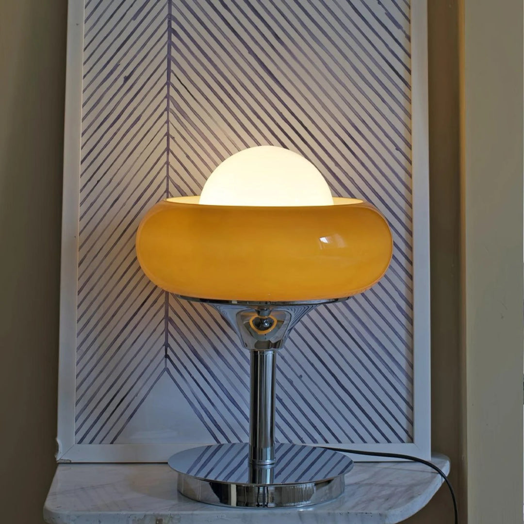 Crostata Table Lamp with a glowing white orb inside a bold orange glass bowl, placed on a marble surface against a decorative striped panel, adding a modern accent to the room with its sleek chrome base and warm illumination.