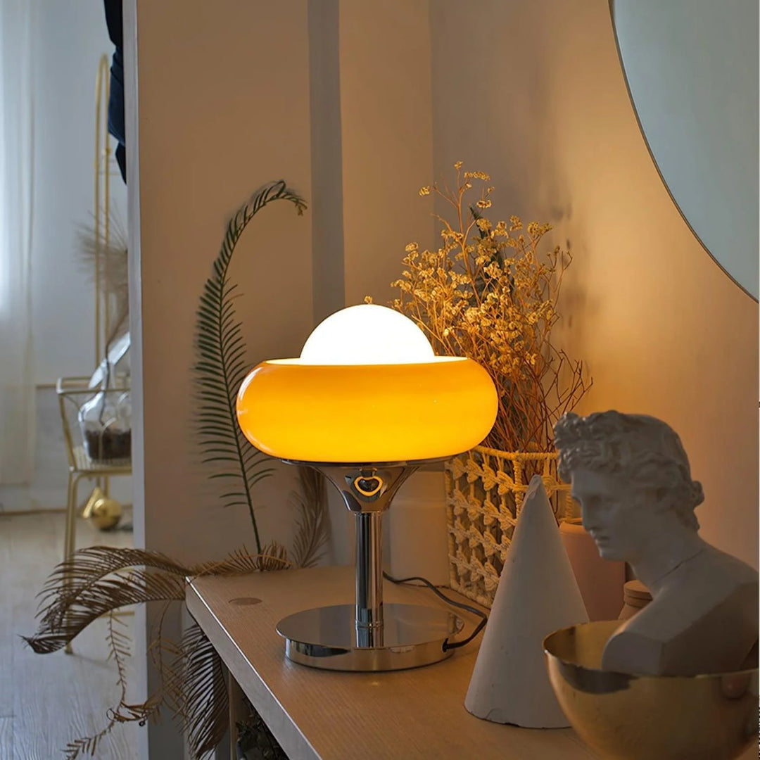 Crostata Table Lamp with a glowing white orb and orange glass bowl, placed on a wooden console table with decorative elements like dried flowers, a sculpture, and a mirror, creating a warm and artistic ambiance in a modern interior.