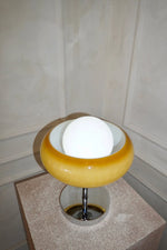 Load image into Gallery viewer, Top-angle view of the Crostata Table Lamp highlighting its glowing white orb nestled inside a vibrant orange glass bowl, displayed on a textured pedestal with a reflective chrome base, set against a neutral wall.
