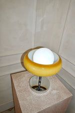 Load image into Gallery viewer, Top view of the Crostata Table Lamp showcasing its spherical glowing orb nestled within a bold orange glass bowl, positioned on a textured pedestal against a neutral-toned wall.
