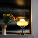 Load image into Gallery viewer, Crostata Table Lamp with a glowing white orb and orange glass bowl, placed on a windowsill beside a vase of vibrant flowers, casting a warm glow against dark blinds, creating a cozy and stylish ambiance.
