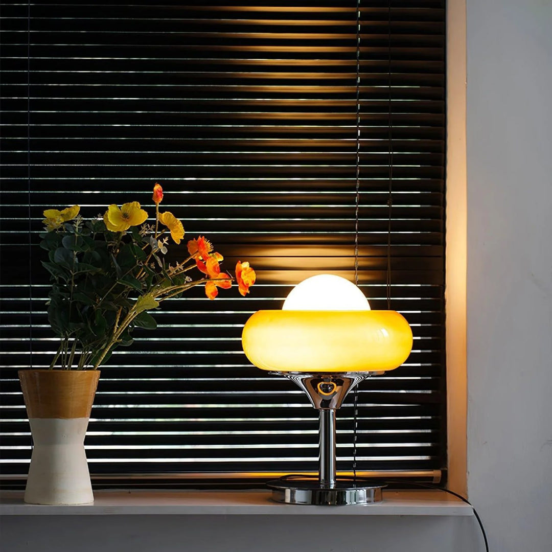 Crostata Table Lamp with a glowing white orb and orange glass bowl, placed on a windowsill beside a vase of vibrant flowers, casting a warm glow against dark blinds, creating a cozy and stylish ambiance.