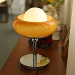 Load image into Gallery viewer, Crostata Table Lamp with an orange glass bowl design and glowing central orb, placed on a wooden table, adding a warm, ambient glow to a modern interior.
