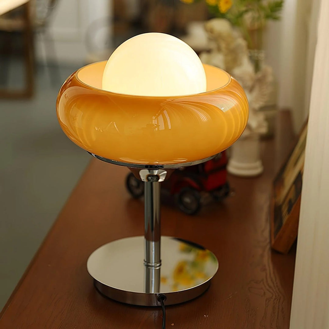 Crostata Table Lamp with an orange glass bowl design and glowing central orb, placed on a wooden table, adding a warm, ambient glow to a modern interior.