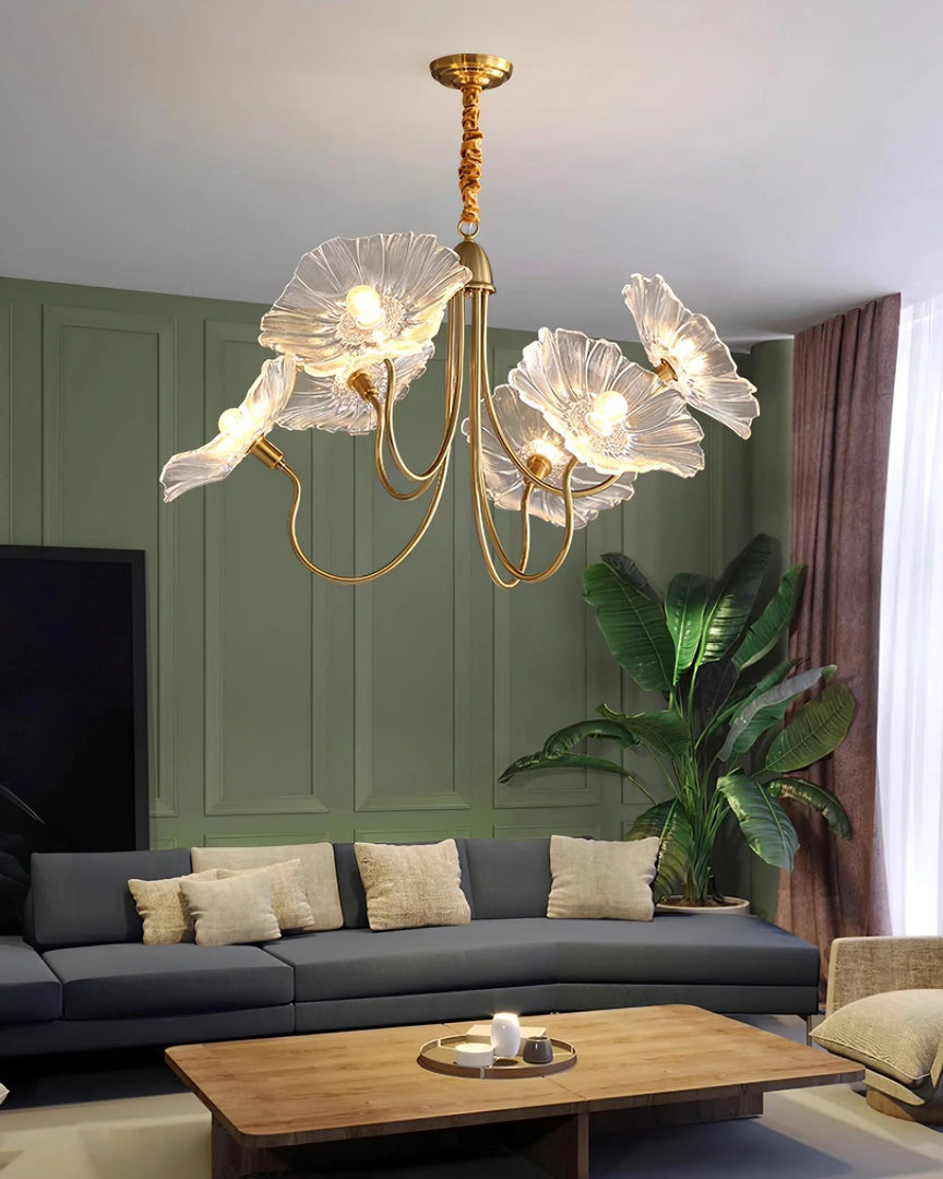 A luxurious crystal floral chandelier with glass petal shades and a gold frame illuminates an elegant living room, featuring a green paneled accent wall, a modern gray sectional sofa with cream cushions, a wooden coffee table, and lush greenery for a serene, upscale ambiance.