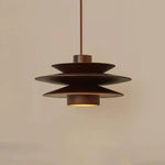 Load image into Gallery viewer, Cylindrical Pendant Light with a layered metal design in a rich brown finish, suspended against a plain beige background, highlighting its sleek form and soft, focused downlight glow.
