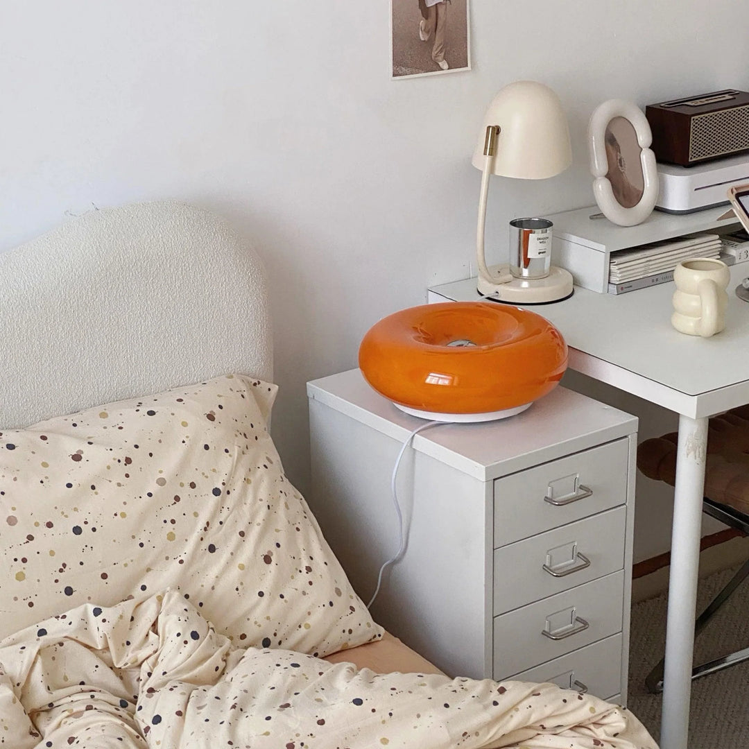 Donuts Table Lamp with a glossy orange ring-shaped design, placed on a bedside cabinet in a cozy bedroom setup featuring soft, neutral bedding, a modern desk lamp, and minimalist decor for a warm and inviting atmosphere.