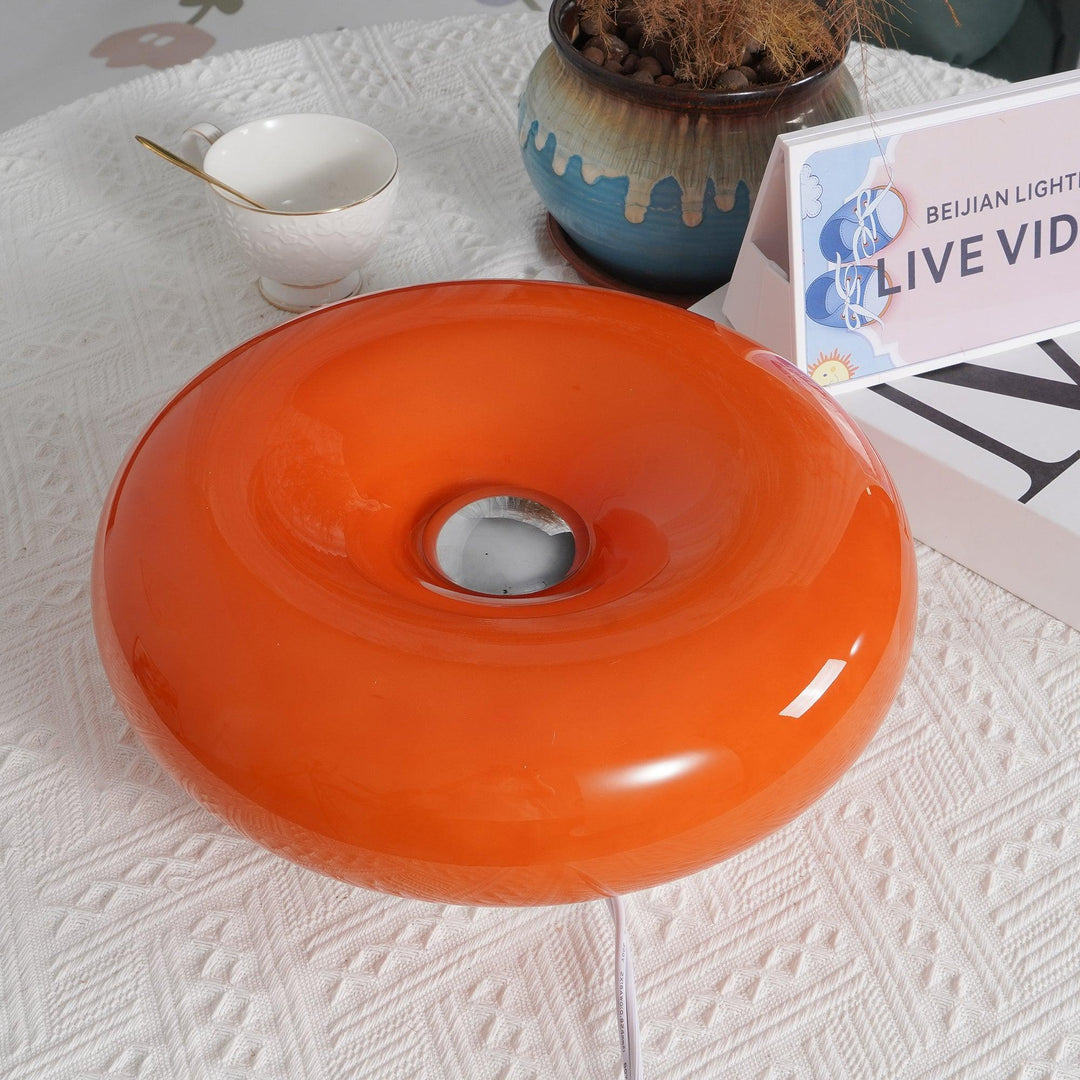 Close-up view of the Donuts Table Lamp showcasing its glossy orange ring-shaped design with a central chrome accent, placed on a textured white surface alongside decorative items such as a teacup and ceramic vase.