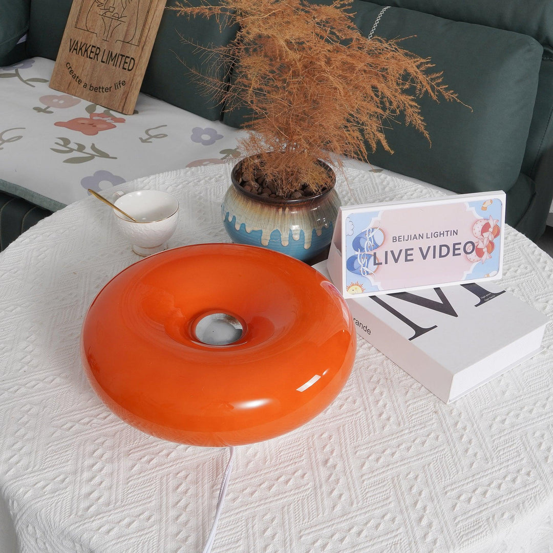 Donuts Table Lamp with a glossy orange ring-shaped design, placed on a textured white tablecloth alongside decorative items, including a ceramic vase, a teacup, and styled with a cozy floral backdrop, creating a charming and inviting setup.