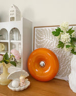 Load image into Gallery viewer, Donuts Table Lamp with a glossy orange ring-shaped design, styled on a wooden surface alongside decorative floral arrangements, ceramic ornaments, and an intricate framed art piece, creating a bright and elegant decorative setup.
