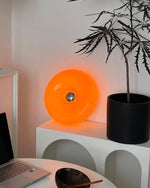 Load image into Gallery viewer, Donuts Table Lamp illuminated with a vibrant orange glow, placed on a modern white arched shelf beside a black planter and minimal decor, creating a cozy and stylish workspace ambiance.
