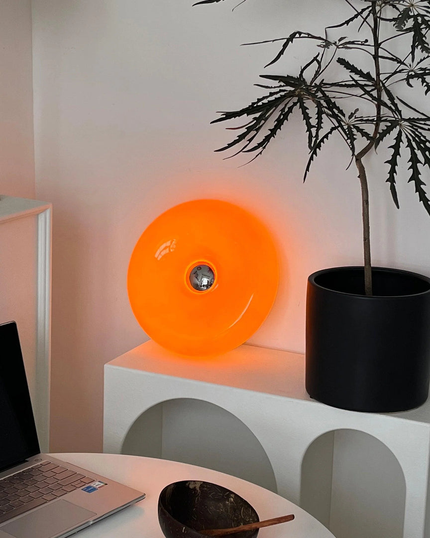 Donuts Table Lamp illuminated with a vibrant orange glow, placed on a modern white arched shelf beside a black planter and minimal decor, creating a cozy and stylish workspace ambiance.