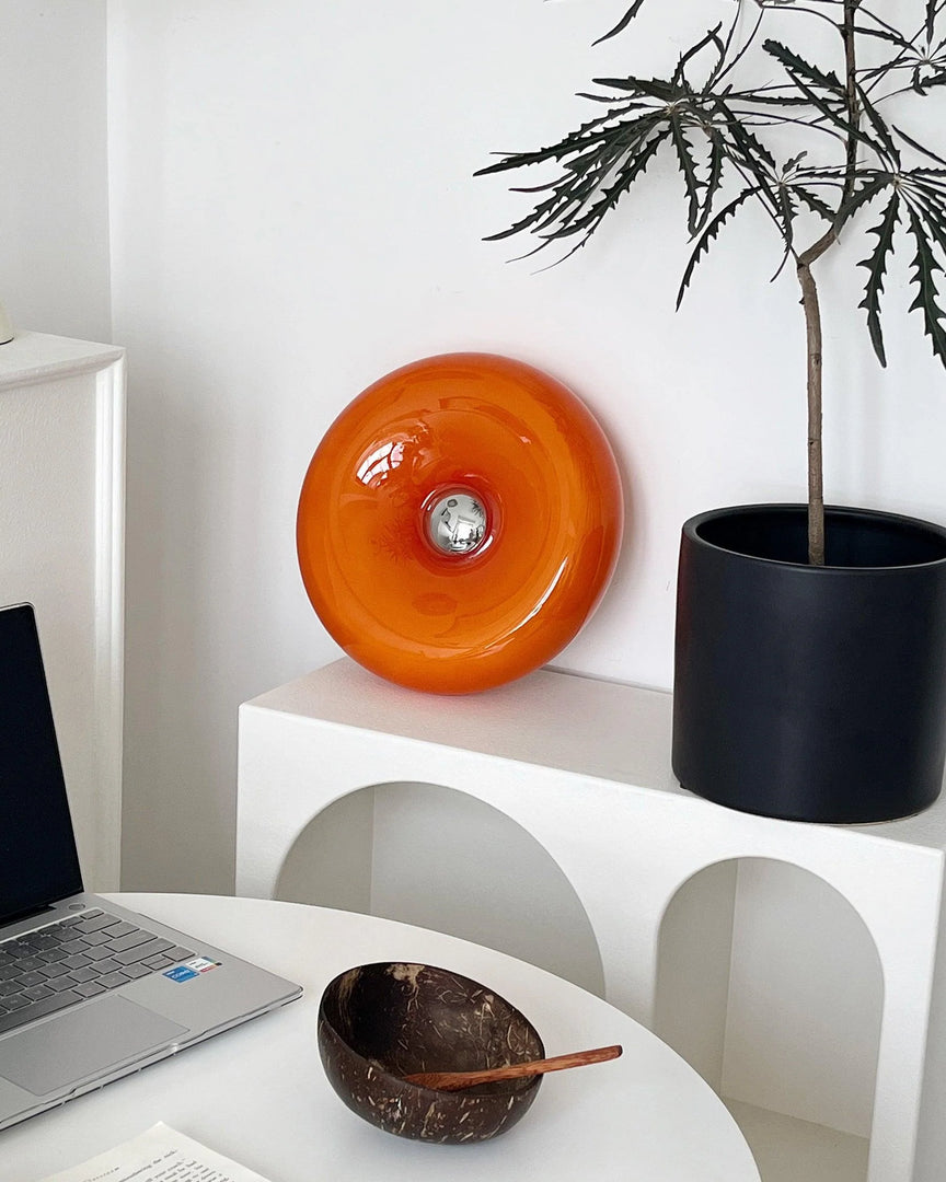 Donuts Table Lamp with a glossy orange ring-shaped design, displayed on a white arched shelf beside minimalist decor, including a black planter and wooden bowl, creating a stylish and modern workspace ambiance.