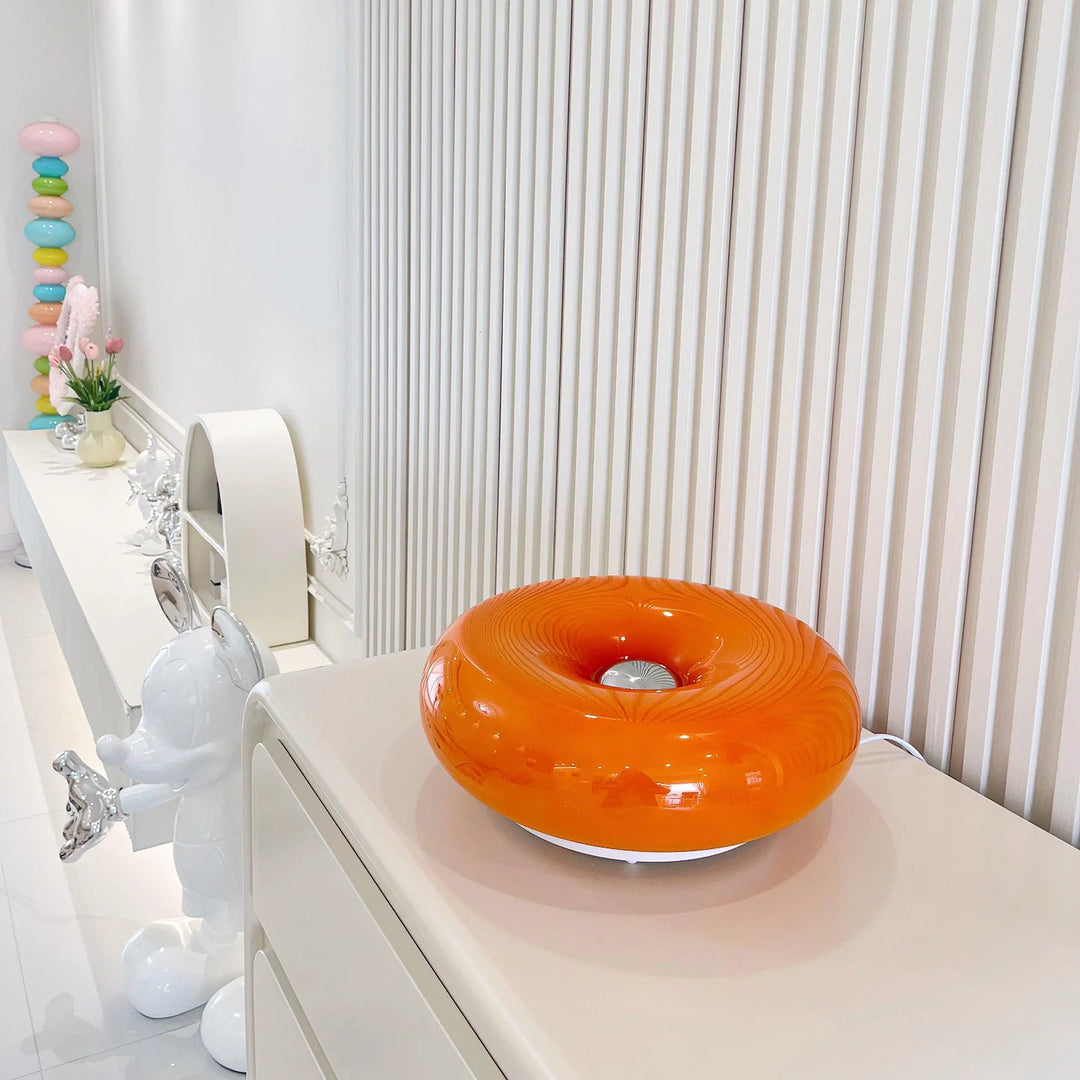 Donuts Table Lamp with a glossy orange ring-shaped design, elegantly placed on a minimalist white console table, surrounded by whimsical decor and set against a backdrop of vertical paneling for a contemporary and playful aesthetic.