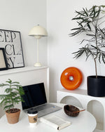 Load image into Gallery viewer, Donuts Table Lamp with a vibrant orange ring-shaped design, displayed in a modern workspace featuring a sleek white desk, neutral decor, a laptop, and coffee, complemented by greenery and framed artwork for a stylish and productive environment.
