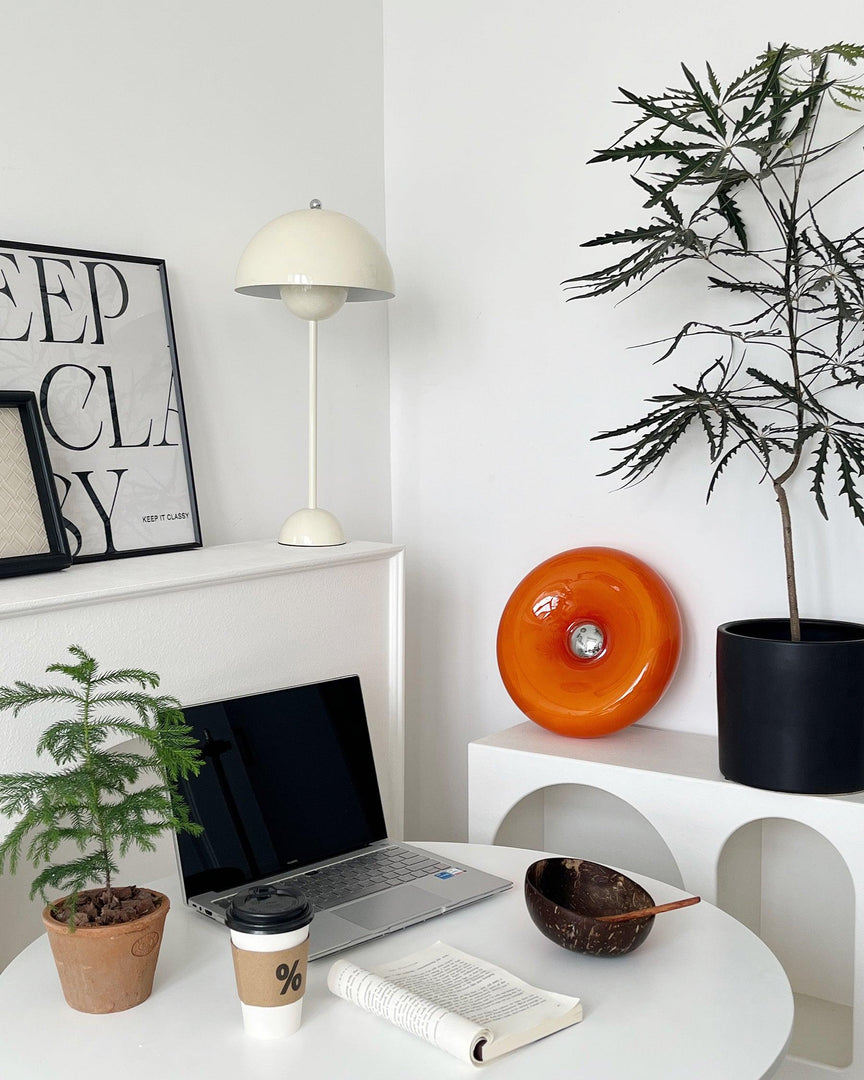 Donuts Table Lamp with a vibrant orange ring-shaped design, displayed in a modern workspace featuring a sleek white desk, neutral decor, a laptop, and coffee, complemented by greenery and framed artwork for a stylish and productive environment.