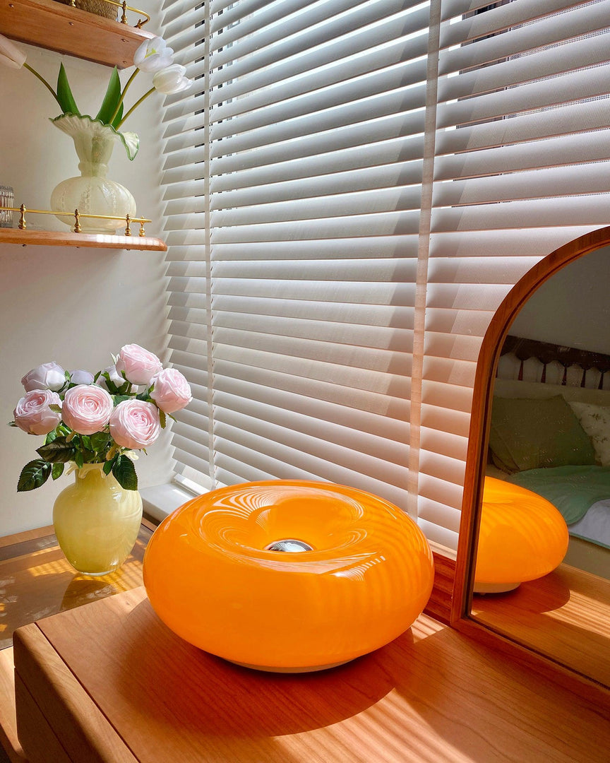 Donuts Table Lamp with a glossy orange ring-shaped design, placed on a wooden dresser reflecting sunlight through window blinds, styled with fresh pink roses in a vase and surrounded by elegant decor for a serene and cozy setting.