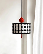 Load image into Gallery viewer, Gracefully designed Metal Lattice Pendant Lamp with a black and white circular motif and vivid red accents, hung near a window draped with a white curtain, enhancing the soft, natural light in a serene setting.
