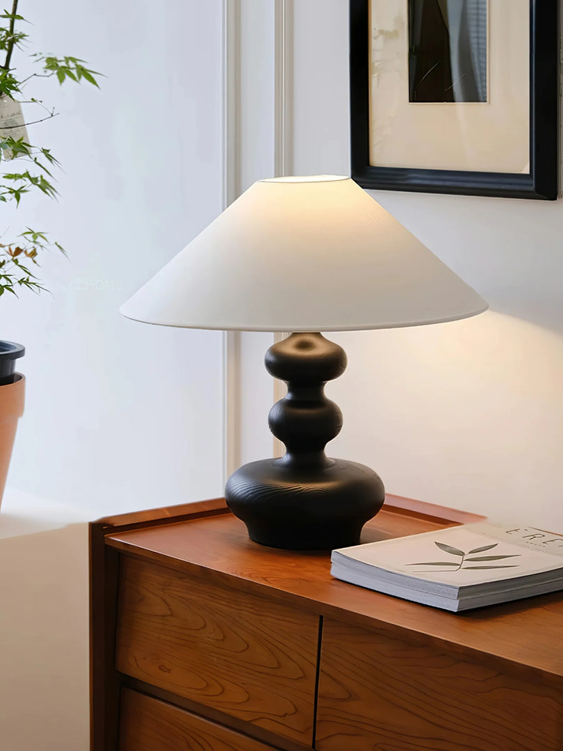 An Elegant Wooden Gourd Lamp gracefully placed on a wooden dresser, featuring a sculpted dark wooden base and a classic white shade. The soft, diffused light complements the peaceful corner of a room adorned with a framed artwork and fresh greenery, creating a refined and tranquil home environment.