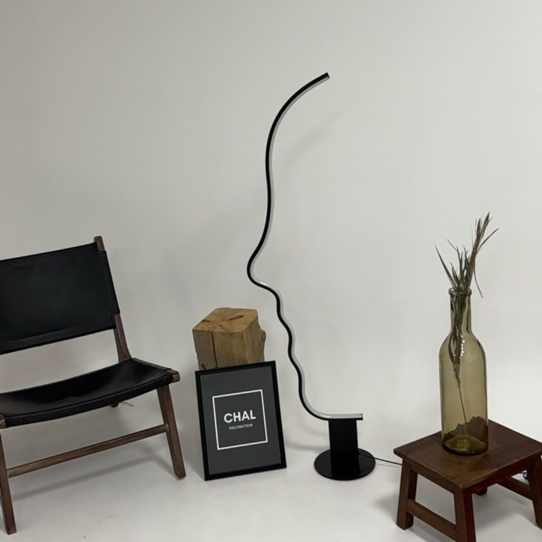 Artistic face-shaped floor lamp in a contemporary display setting with a black chair, decorative wood accents, a framed art piece, and a glass vase on a small stool.