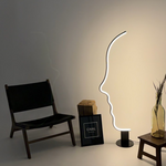 Load image into Gallery viewer, Illuminated face-shaped floor lamp casting a warm ambient glow, styled with a black chair, wooden accents, framed artwork, and a decorative glass vase in a modern setting.
