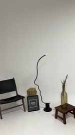 Load image into Gallery viewer, Minimalist room display featuring a face-shaped lamp with a sleek black frame, styled alongside a black chair, a decorative wooden block, framed artwork, and a glass vase with dried stems on a wooden stool.
