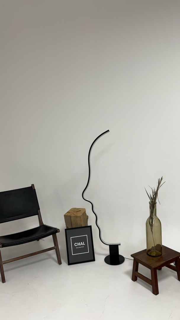 Minimalist room display featuring a face-shaped lamp with a sleek black frame, styled alongside a black chair, a decorative wooden block, framed artwork, and a glass vase with dried stems on a wooden stool.