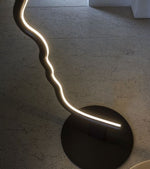 Load image into Gallery viewer, Side-angle view of the face-shaped lamp showcasing its smooth wavy contours and illuminated edge, standing on a black circular base against a textured floor.
