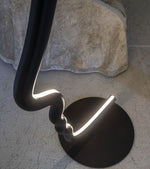 Load image into Gallery viewer, Overhead view of the face-shaped lamp’s elegant curved design and black circular base, highlighting its glowing edges on a light-toned floor near a draped fabric.
