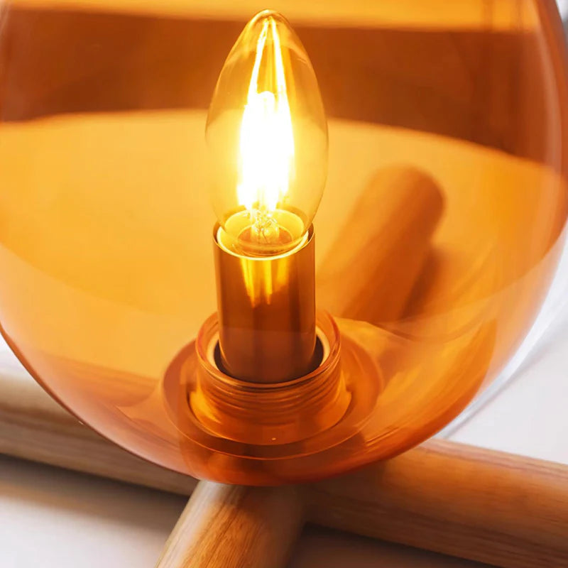 Close-up view of the glowing filament bulb inside the amber glass teardrop-shaped shade of the Firebeam table lamp, highlighting the warm light and intricate wooden cross-base details.