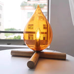 Load image into Gallery viewer, Firebeam table lamp with an amber glass teardrop-shaped shade and glowing bulb, placed on a table near a window with a view of greenery and buildings, adding warmth to the natural light-filled room.
