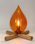 Load image into Gallery viewer, Close-up view of the Firebeam table lamp, showcasing its amber teardrop-shaped glass shade, glowing filament bulb, and natural wooden cross-base, creating a warm ambient lighting effect.
