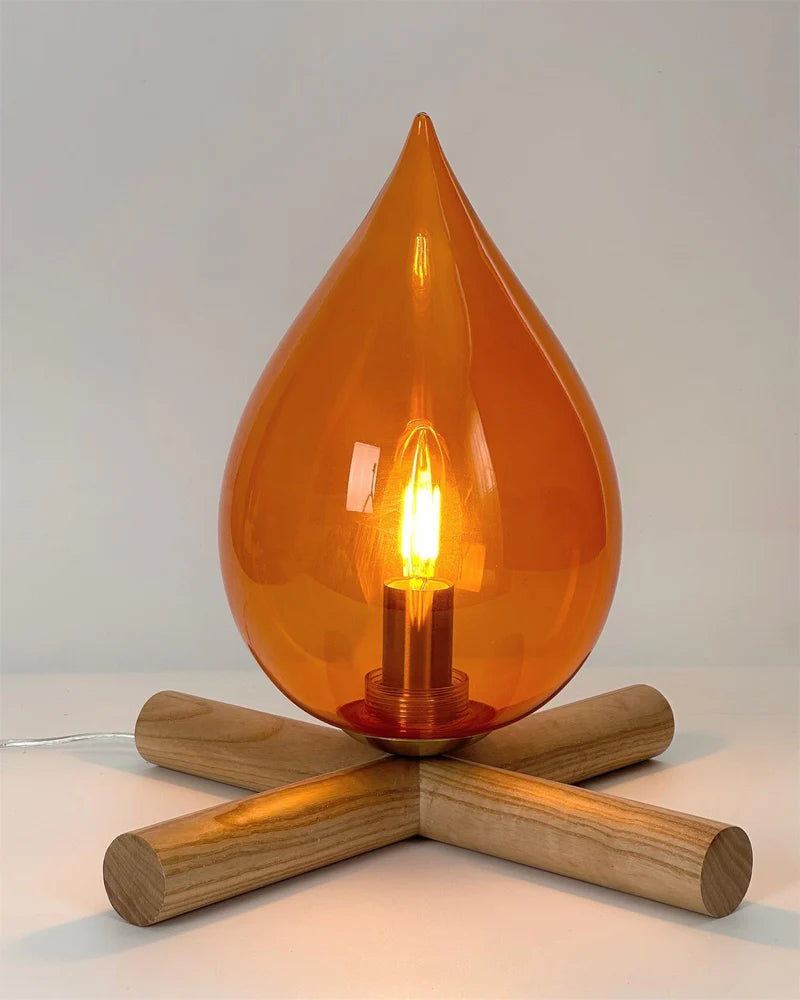 Close-up view of the Firebeam table lamp, showcasing its amber teardrop-shaped glass shade, glowing filament bulb, and natural wooden cross-base, creating a warm ambient lighting effect.