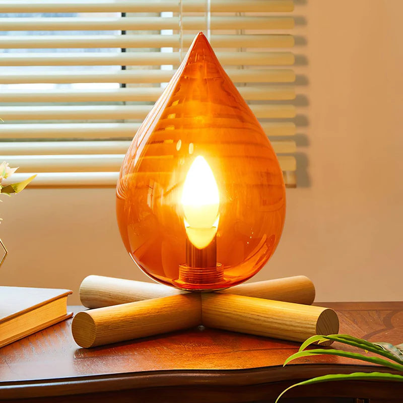 Firebeam table lamp with an amber glass teardrop shade and glowing bulb, placed on a wooden table near a window with blinds, adding a cozy and decorative touch to the space.