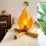 Load image into Gallery viewer, Firebeam table lamp with an amber teardrop-shaped glass shade and glowing bulb, placed on a white glossy table in a modern living room, surrounded by greenery and elegant decor.
