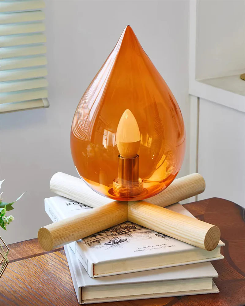 Firebeam table lamp with an amber teardrop-shaped glass shade placed on stacked books, featuring a glowing bulb and wooden cross-base, creating a cozy and decorative reading nook ambiance.