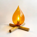 Load image into Gallery viewer, Firebeam table lamp featuring an amber glass teardrop-shaped shade with a warm glowing bulb, resting on a unique wooden cross-base for a cozy and stylish lighting design.
