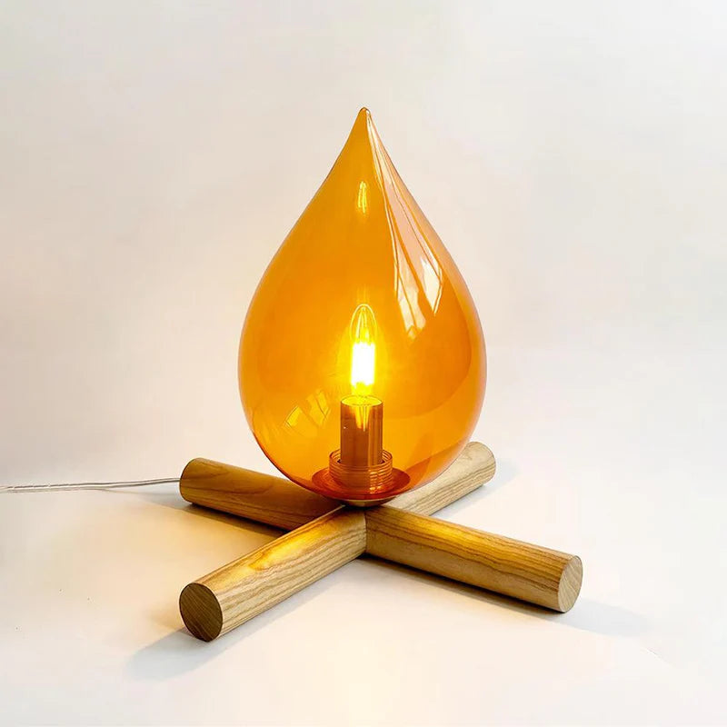 Firebeam table lamp featuring an amber glass teardrop-shaped shade with a warm glowing bulb, resting on a unique wooden cross-base for a cozy and stylish lighting design.