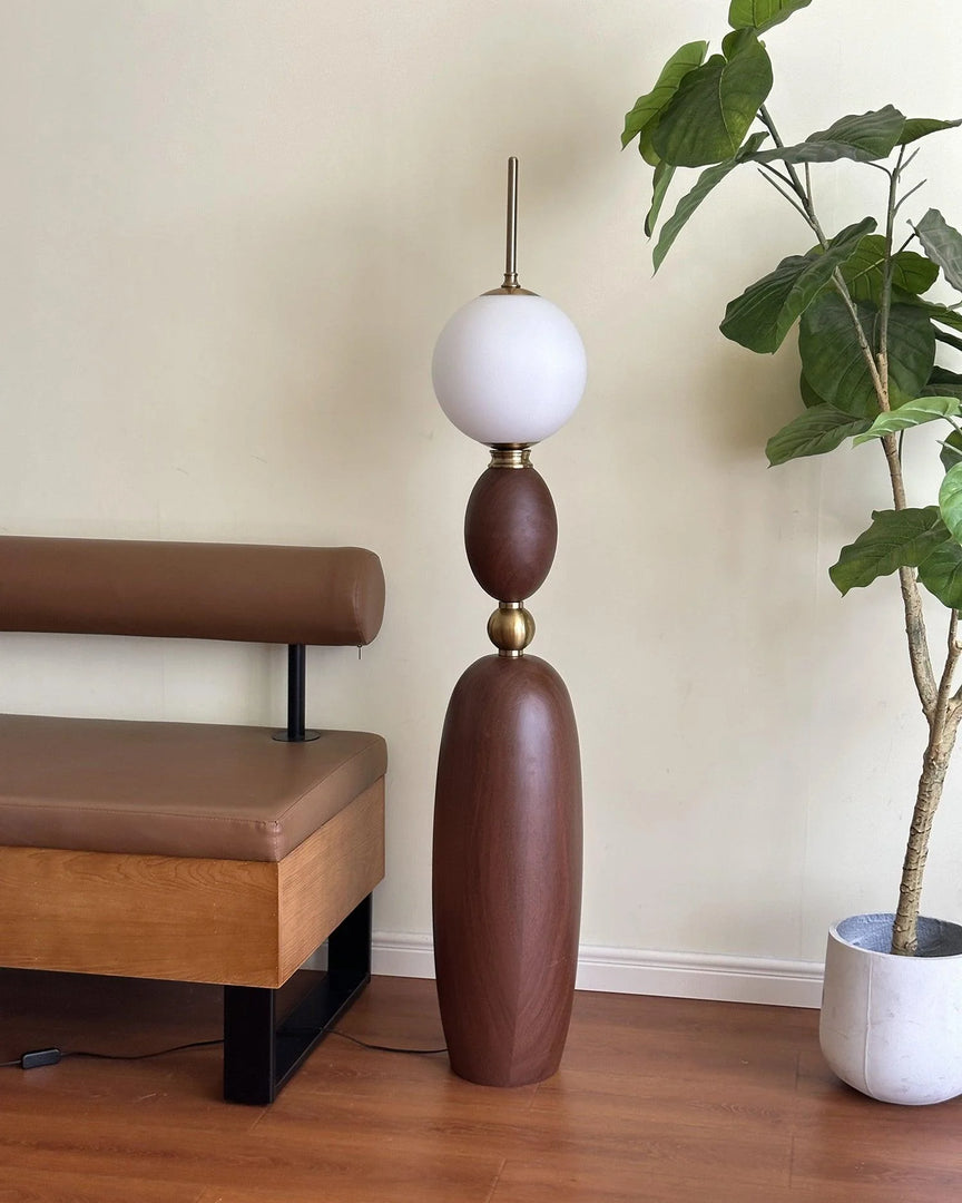 Layali floor lamp with a wooden sculptural base, gold accents, and a frosted globe shade, unlit, placed beside a modern leather bench and a potted indoor plant, creating a warm and inviting corner space.