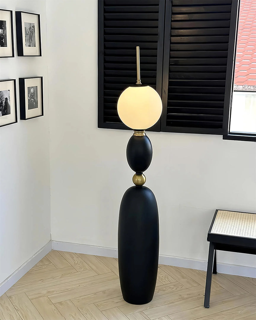 Layali floor lamp with a black sculptural base, gold accents, and a glowing frosted globe shade, placed in a modern corner with black shutters, framed monochrome wall art, and a minimalist bench.