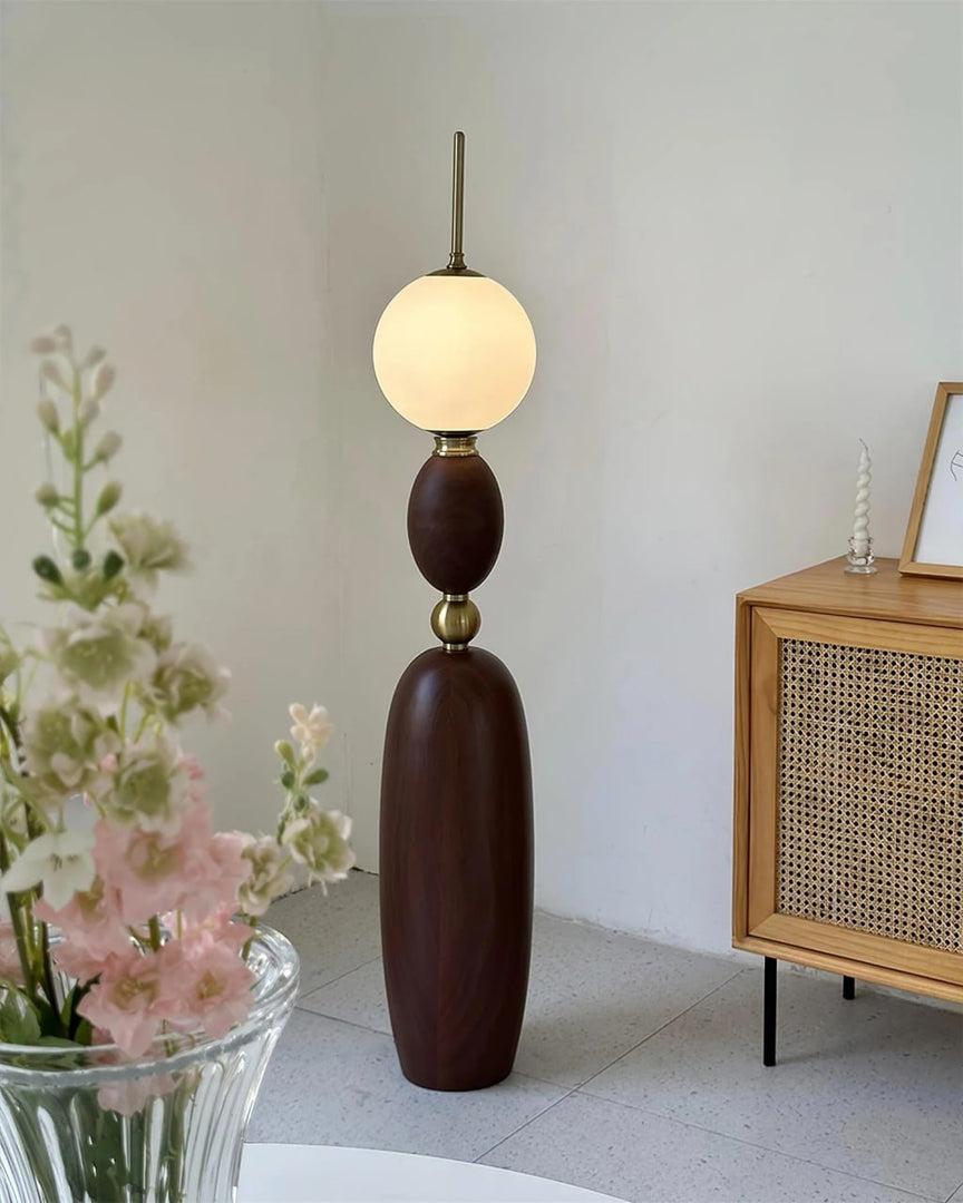 Layali floor lamp with a sculptural wooden base, gold accents, and a glowing frosted globe shade, placed in a bright modern room with a rattan-front cabinet and floral centerpiece for a soft and elegant ambiance.