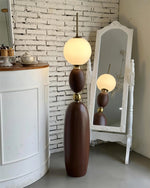 Load image into Gallery viewer, Layali floor lamp with a wooden sculptural base, gold accents, and a glowing frosted globe shade, styled in front of a rustic white brick wall beside a decorative white-framed mirror and a vintage counter.
