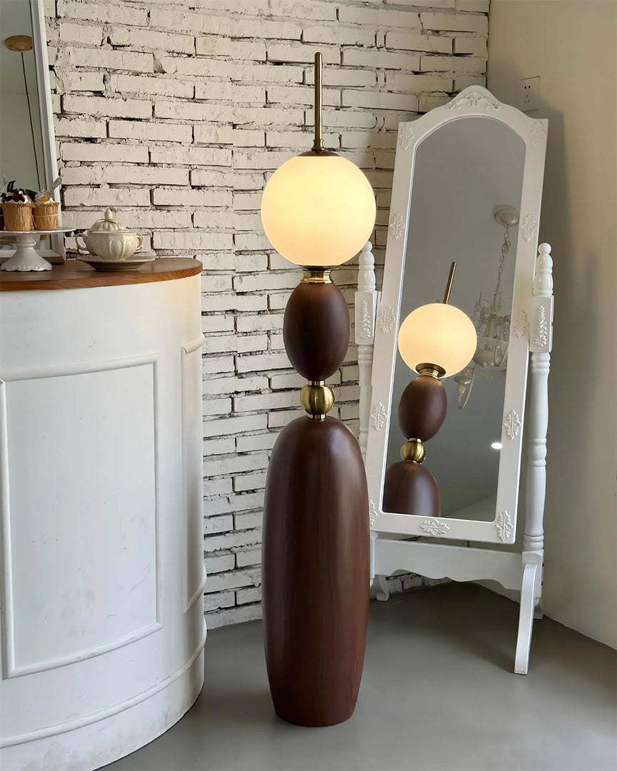 Layali floor lamp with a wooden sculptural base, gold accents, and a glowing frosted globe shade, styled in front of a rustic white brick wall beside a decorative white-framed mirror and a vintage counter.