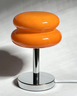 Load image into Gallery viewer, A stylish Glossy Macaron Table Lamp featuring two stacked, glossy orange discs on a sleek chrome stand with a round base, illuminated against a shadowed background.
