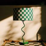 Load image into Gallery viewer, Featuring the Green Dama Lamp, this image captures a striking green and white checkered lampshade atop a spiraled, vibrant green base. The unique, playful design casts intriguing shadows and highlights, set against a warm, sunlit background, reflecting the lamp&#39;s dynamic and modern aesthetic. Perfect for adding a bold statement to any room.
