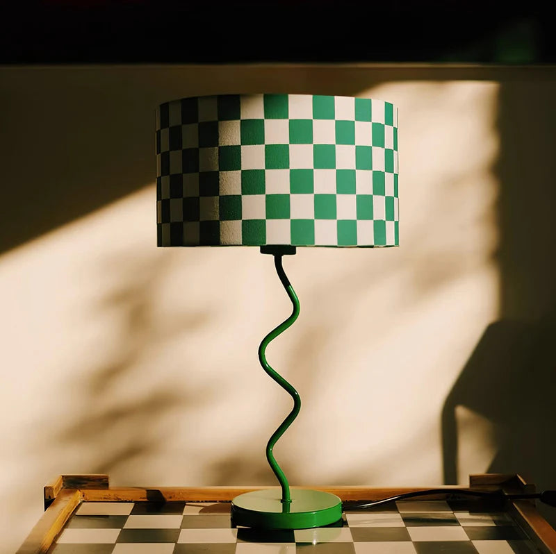 Featuring the Green Dama Lamp, this image captures a striking green and white checkered lampshade atop a spiraled, vibrant green base. The unique, playful design casts intriguing shadows and highlights, set against a warm, sunlit background, reflecting the lamp's dynamic and modern aesthetic. Perfect for adding a bold statement to any room.