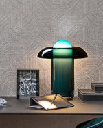 Load image into Gallery viewer, Green glass table lamp with a glowing dome placed on a modern desk, styled with a geometric-patterned wallpaper, a monochrome photo frame, a black leather organizer, and a pen holder.
