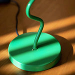 Load image into Gallery viewer, A close-up view of the vibrant green base of a spiral lamp, highlighting its smooth, glossy finish and whimsical twisted stem. The warm sunlight streaming in casts dynamic shadows across the wooden surface, enhancing the sleek and modern design of this playful decor piece.
